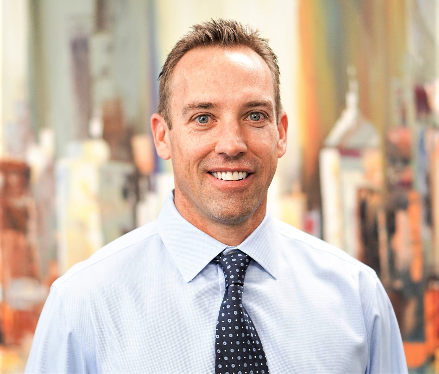 Man wearing a light blue dress shirt and patterned tie, smiling at the camera, with an abstract blurred background.