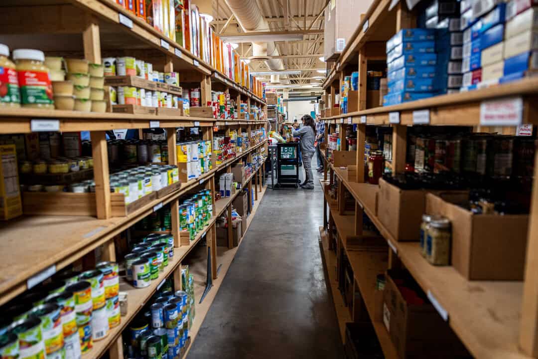 1S9A3608 – Nora Two people with a cart walk down an aisle lined with shelves stocked with canned and boxed food in a warehouse or food pantry.