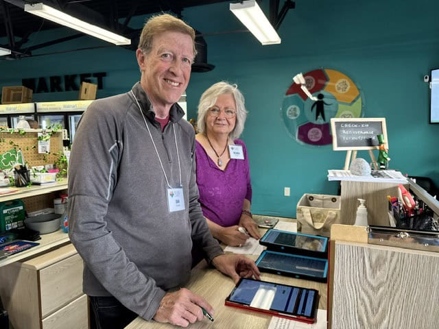 IMG_5098 – Nora Two people stand at a counter with several tablets, both wearing name tags and smiling, in a room with teal walls and a colorful wall graphic.