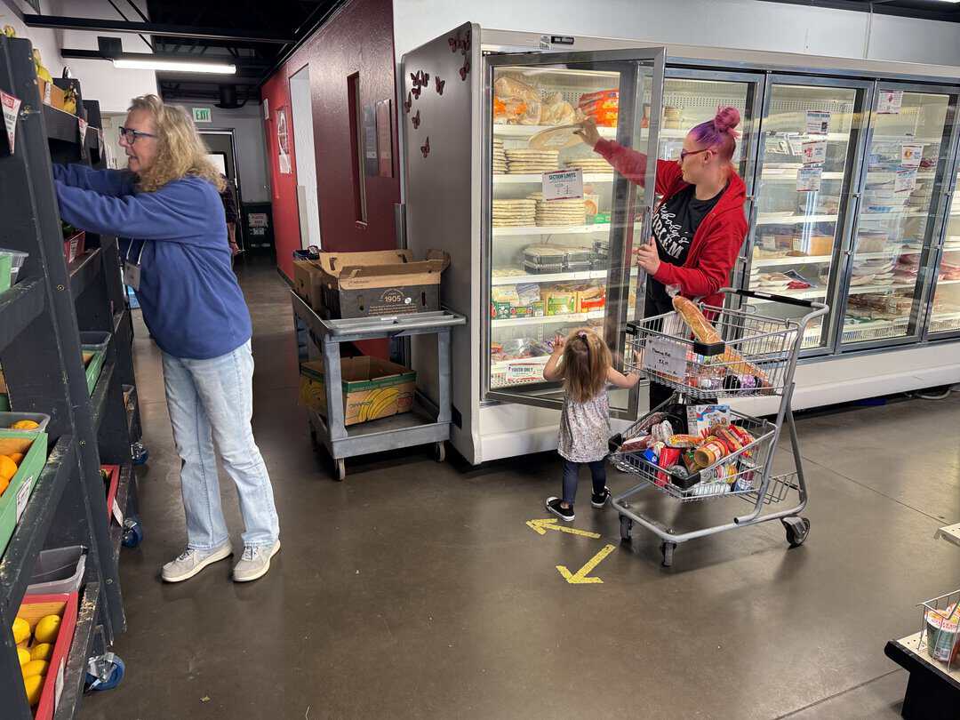 Joy_20260312_152250_959_photo – Nora A woman and child shop for groceries near a refrigerator, while a man selects produce from a shelf in a store.