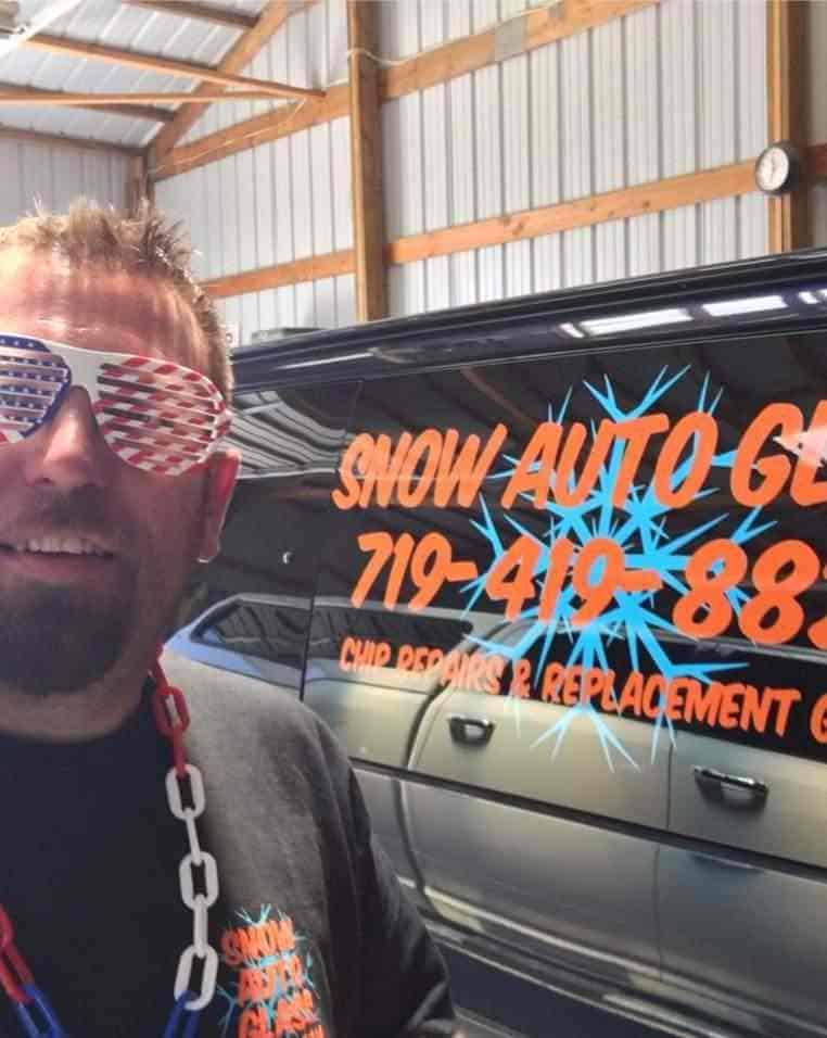 Rian Snow Auto Glass Man wearing American flag sunglasses and a red, white, and blue necklace stands in front of a van with “Snow Auto Glass” and a phone number written on the side.