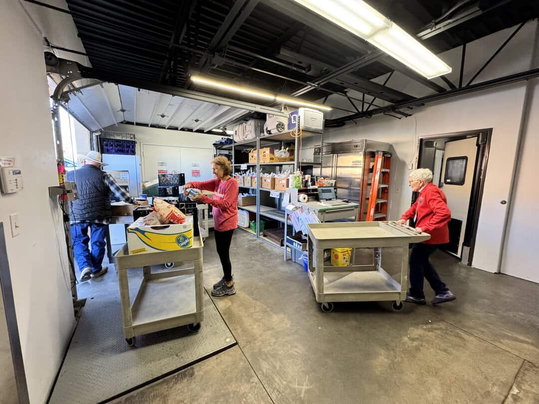 Unloading Stock April 2026 – Nora Three people organize and move food items in a storage room with shelves, carts, and boxes under fluorescent lighting.